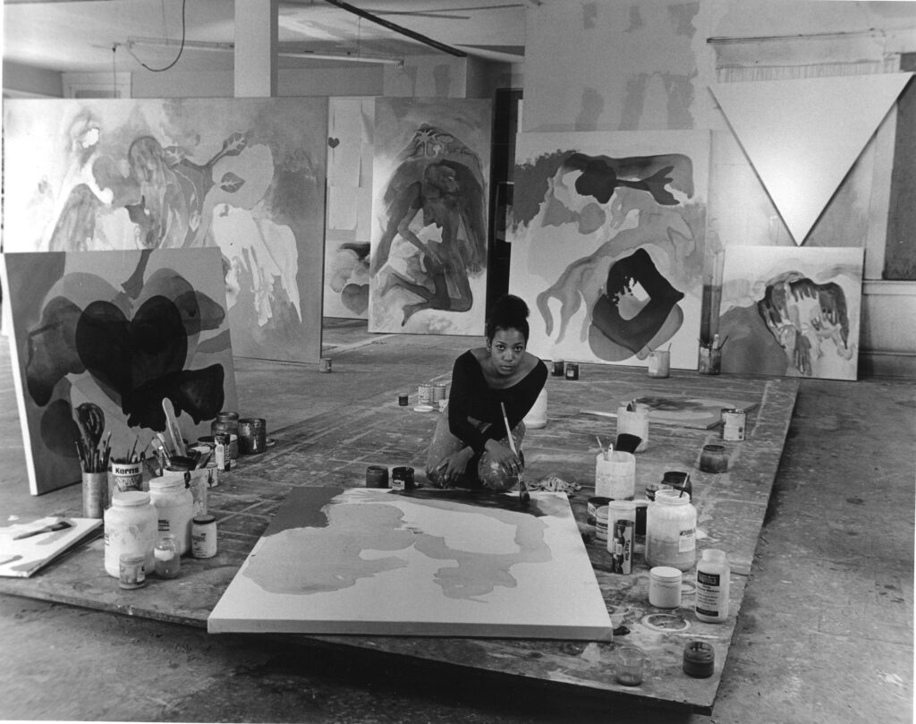 A black and white photograph of artist Suzanne Jackson painting in her studio with large abstract artworks and paint supplies in the background.